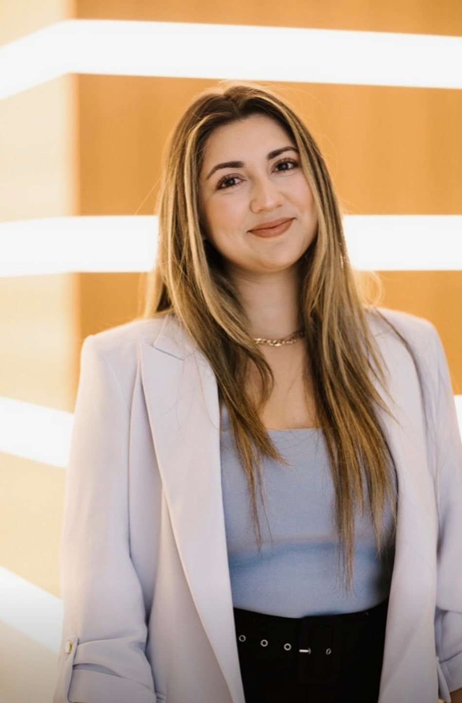 Alexandra Marie in a pale blazer over a light blue top, standing in front of a warm timber wall with horizontal light bands