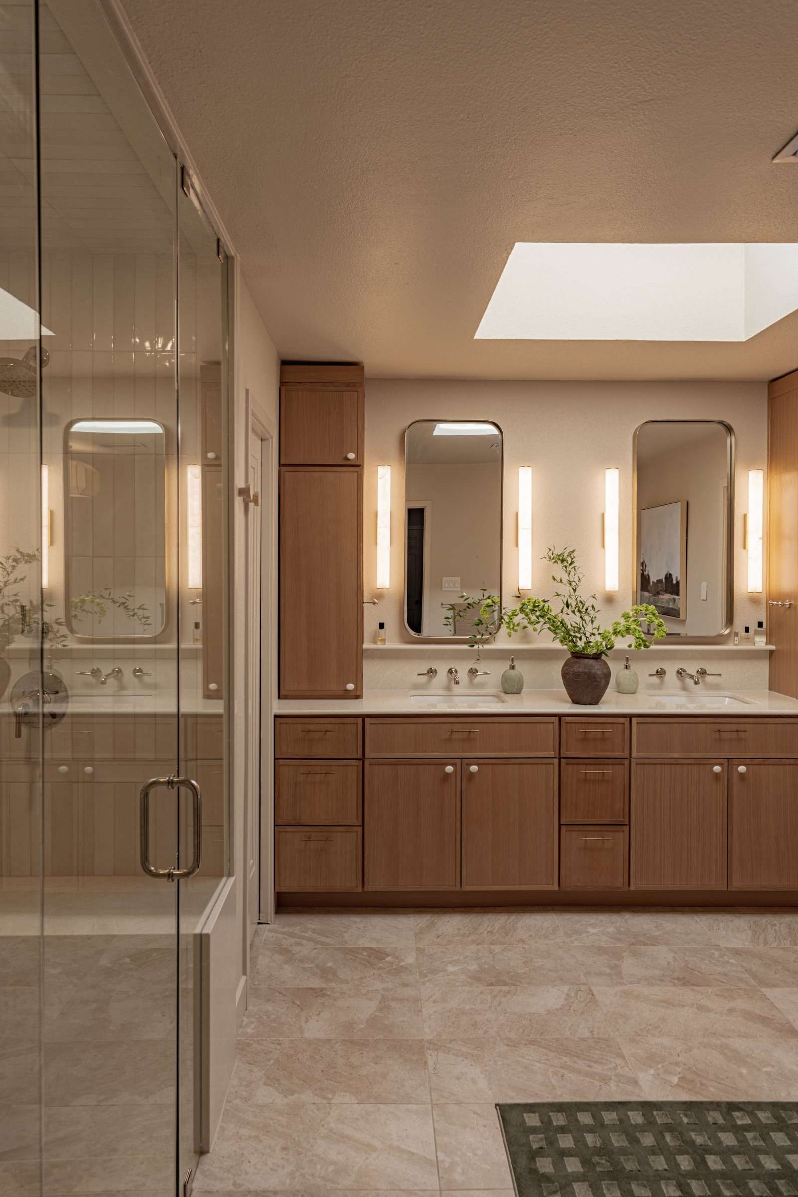 Primary bathroom with a double vanity in white oak under a skylight, twin rounded mirrors between vertical sconces, and a glass shower enclosure to one side on stone-look tile
