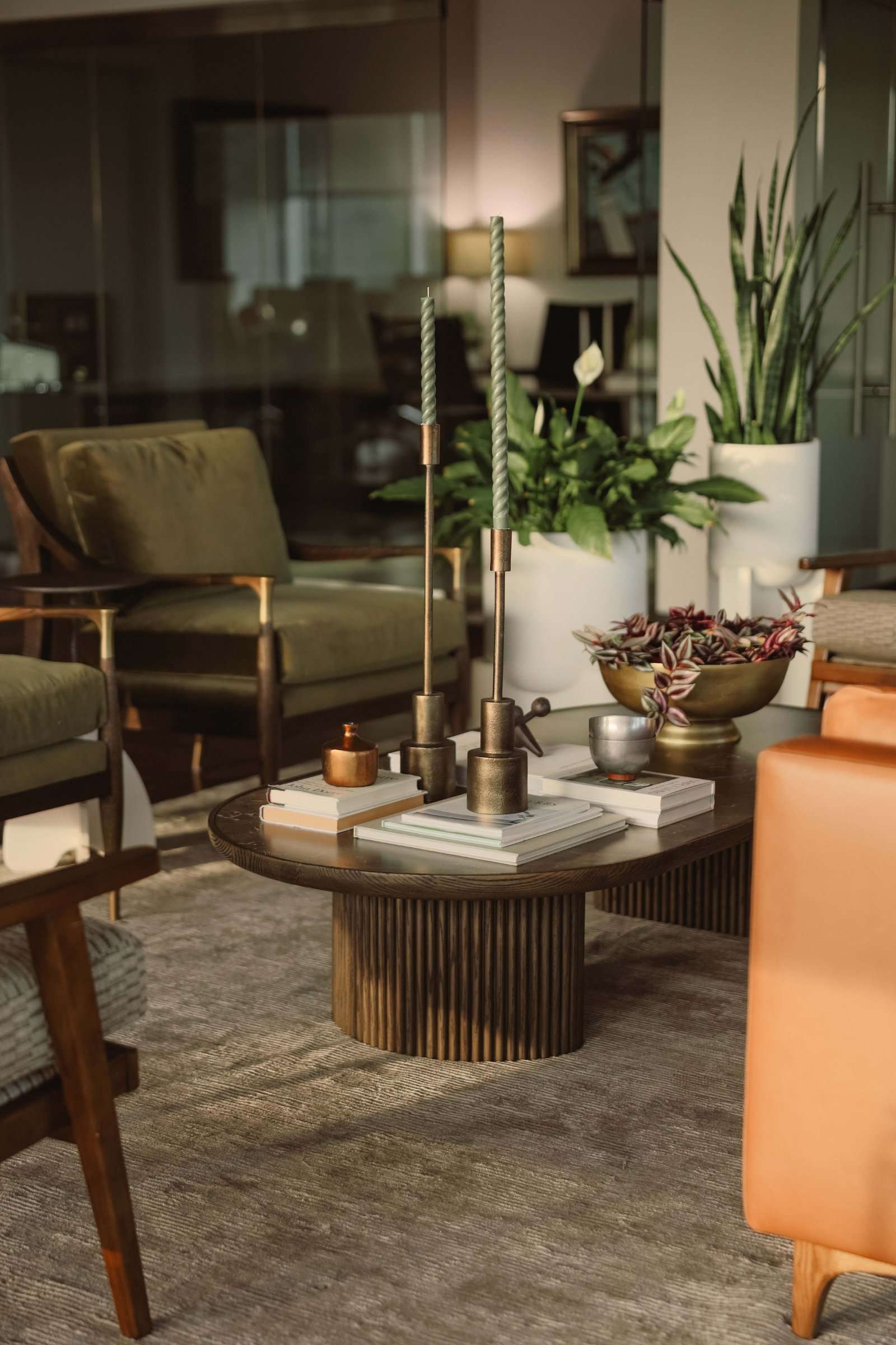 Office lounge: a fluted-base oval table with brass candlesticks and books, olive velvet armchairs behind, a tan leather sofa at the edge of frame and glass partitions beyond