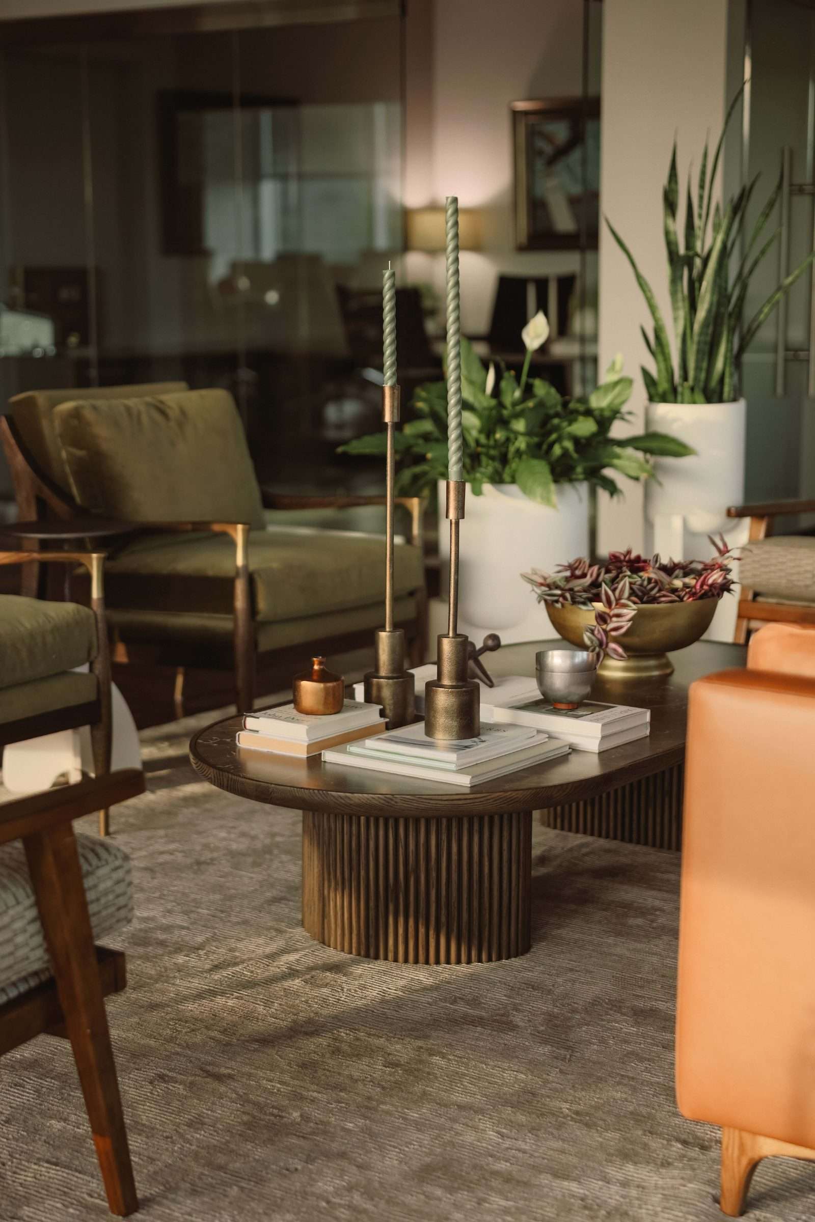 Office lounge: a fluted-base oval table with brass candlesticks and books, olive velvet armchairs behind, a tan leather sofa at the edge of frame and glass partitions beyond