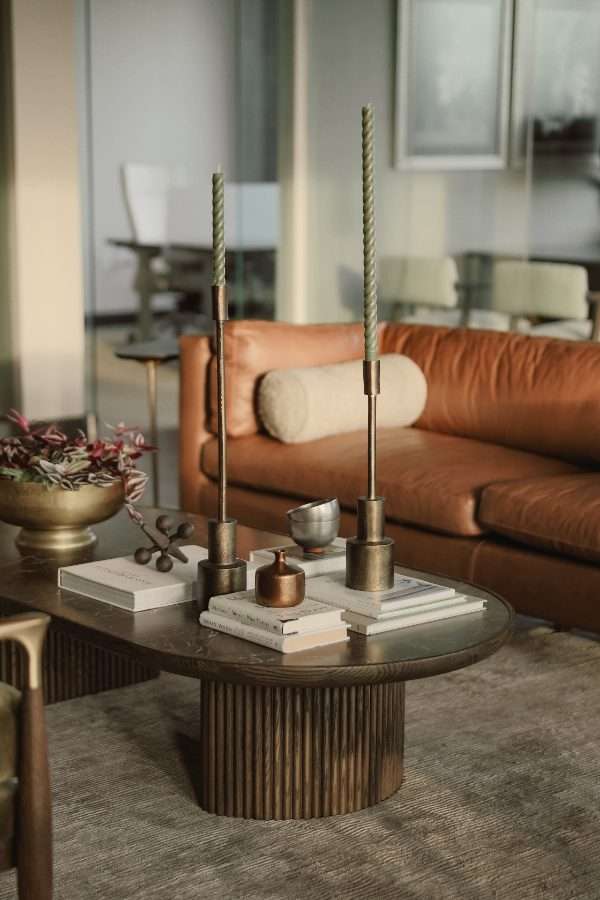 Fluted timber coffee table styled with tall candlesticks, stacked books and a brass bowl, in front of a tan leather sofa with a bolster cushion