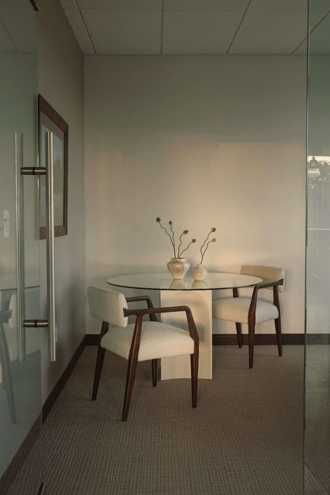 Small meeting corner behind a glass partition: a round glass table on a pale plinth base with two boucle armchairs and dried stems in ceramic vases
