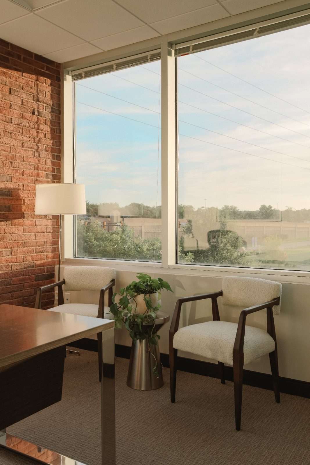Corner office with an exposed brick wall and ribbon windows, two boucle armchairs beside a metal side table with a trailing pothos, and a desk in the foreground