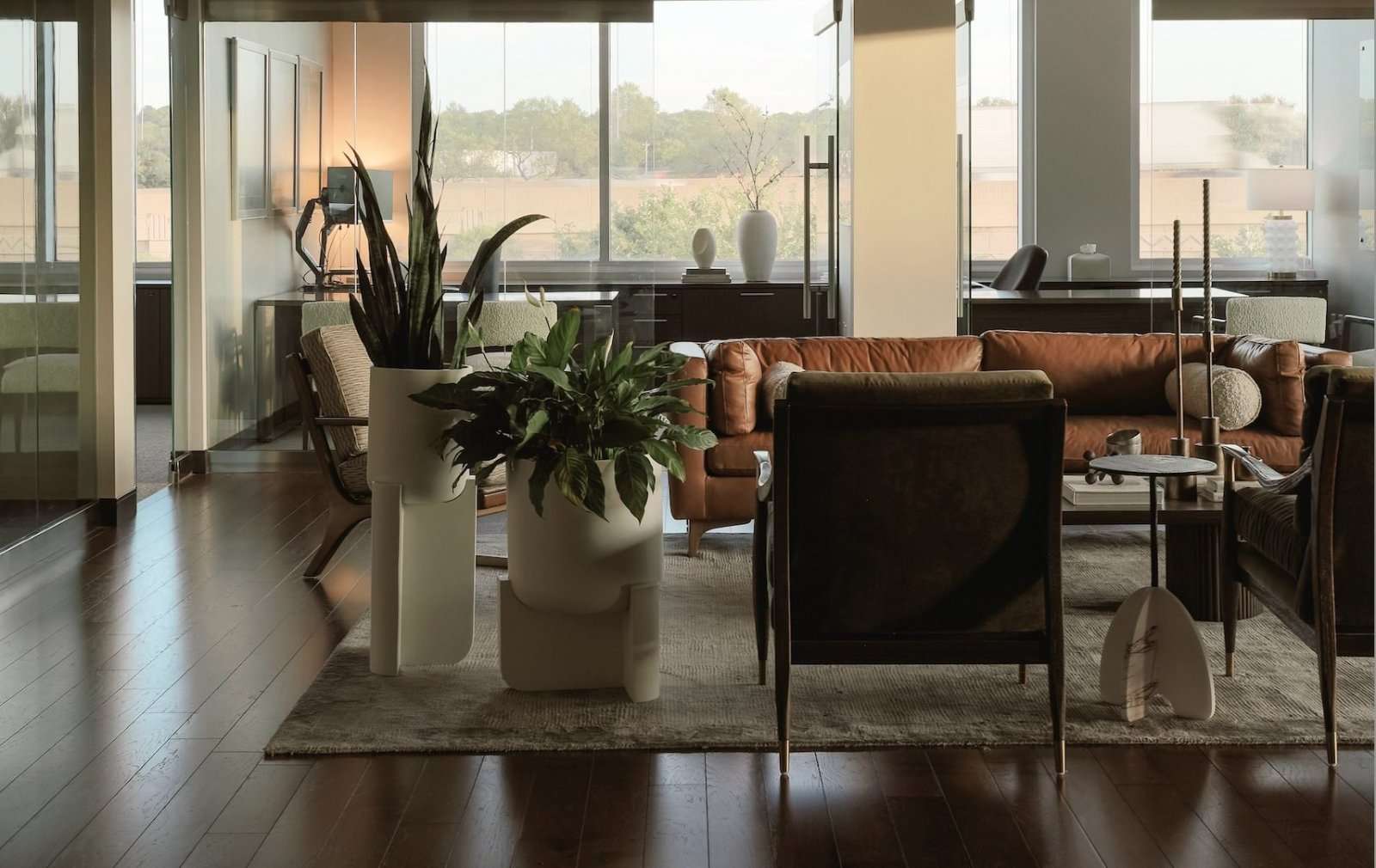 Open office lounge on dark timber floors: a tan leather sofa and dark armchairs around a low table, two large sculptural planters, and glazed offices along the window wall