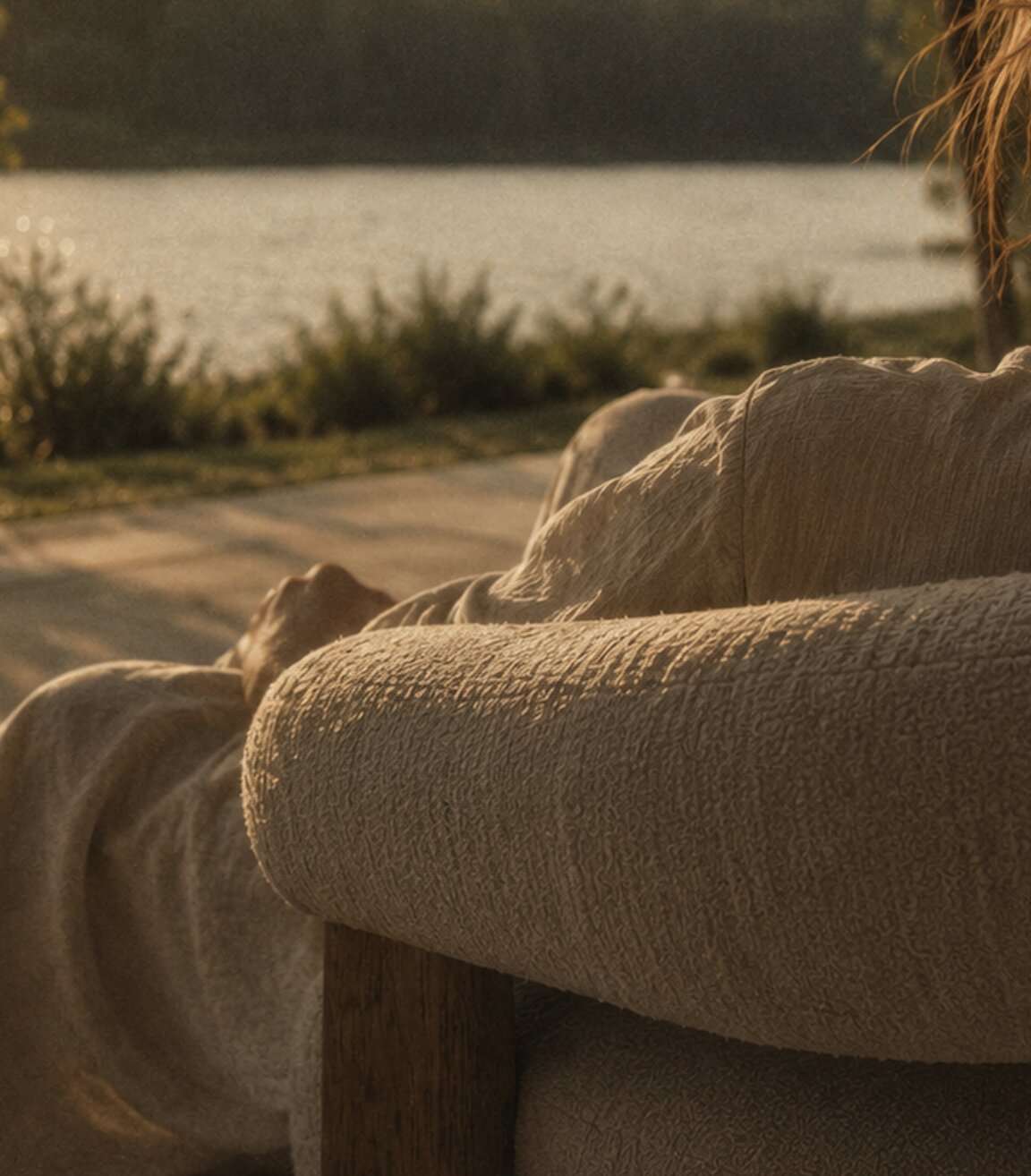 Artist impression — close view over the back of a textured bouclé sofa, someone reclining on it, looking out to open water in low golden light