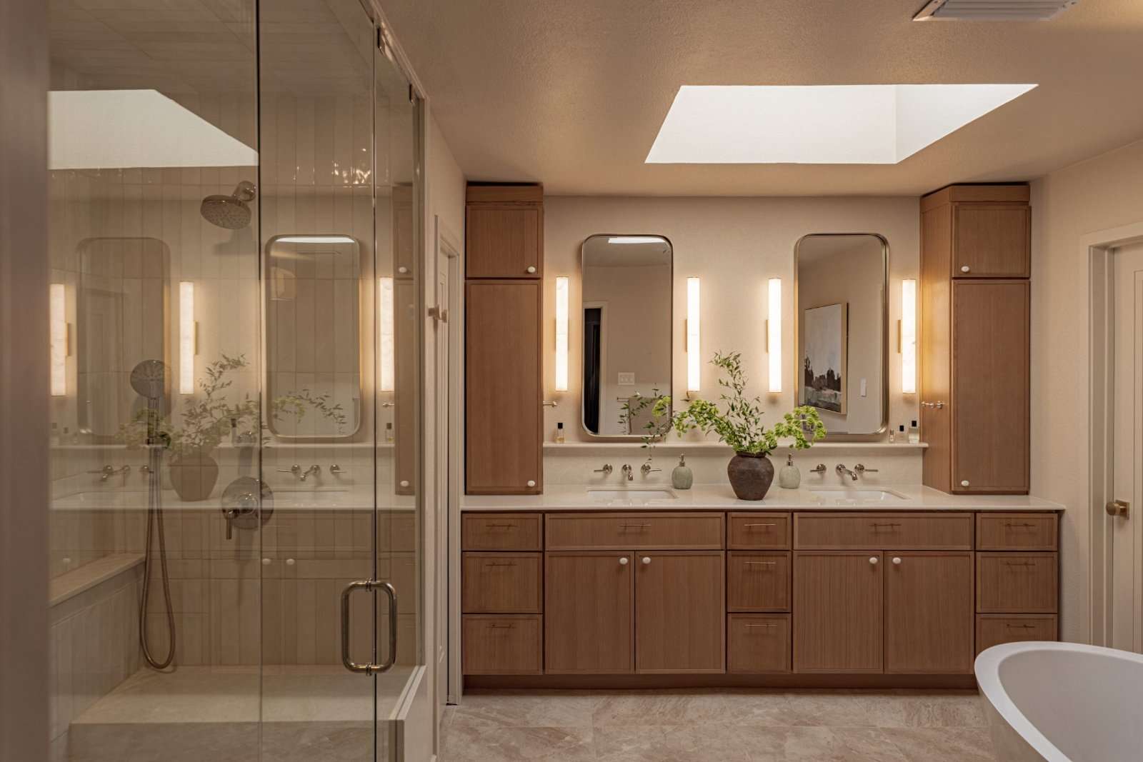 Primary bathroom seen across the room: white oak vanity and full-height storage towers under a skylight, wall-mounted taps, a glazed shower to the left and the edge of a freestanding tub at the right