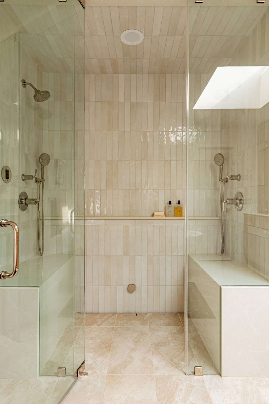 Walk-in steam shower clad in glazed cream tile, with facing stone benches, twin showerheads and handsets, and a full-width niche