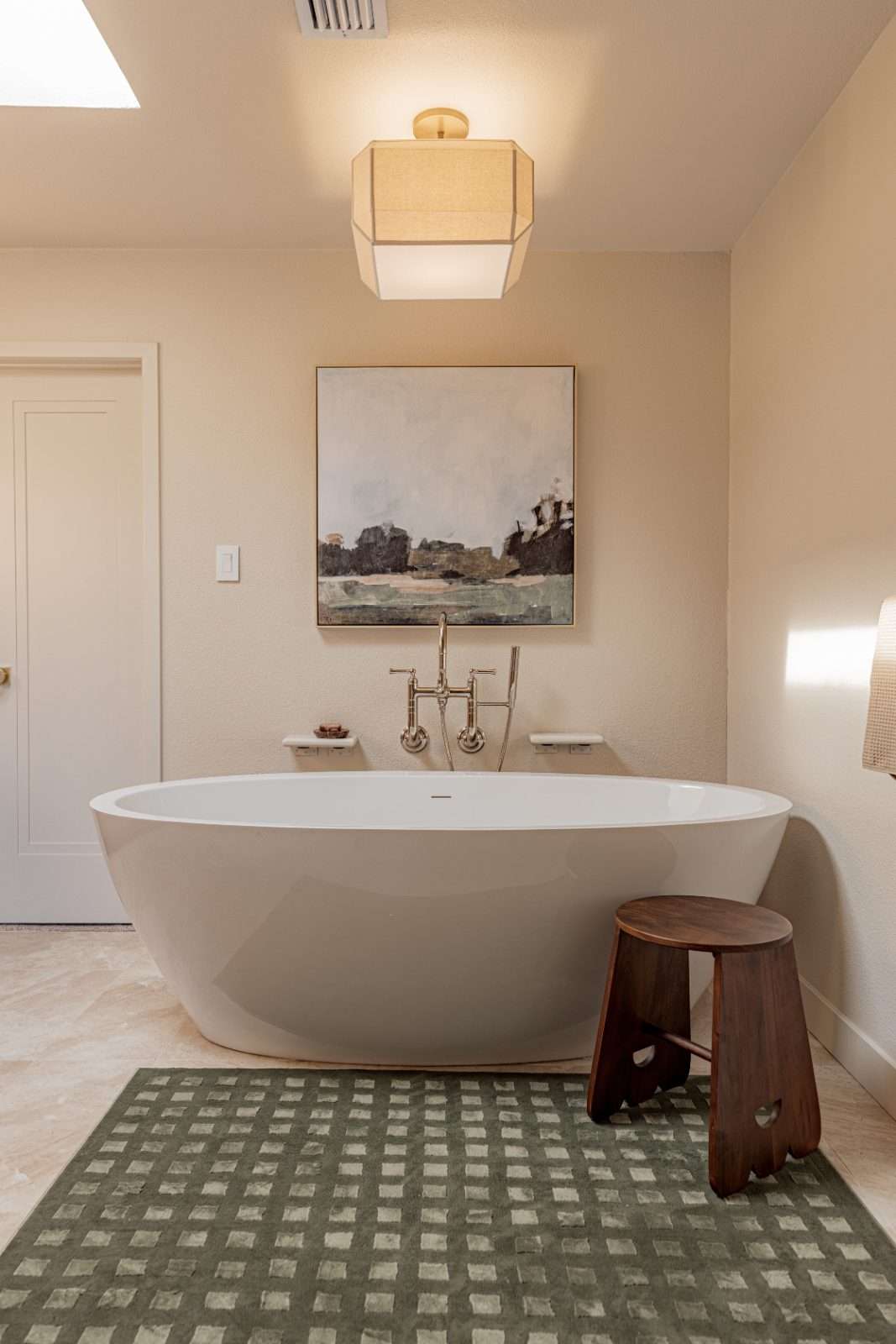 Freestanding oval tub with a floor-mounted tap below a framed landscape painting, a dark timber stool alongside and a green checked rug underfoot