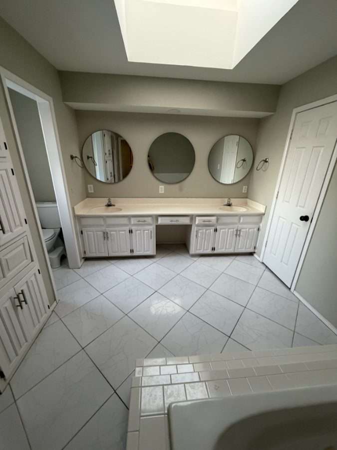 The bathroom before the remodel: white raised-panel cabinets with a laminate counter, three round mirrors, and glossy white marbled floor tile