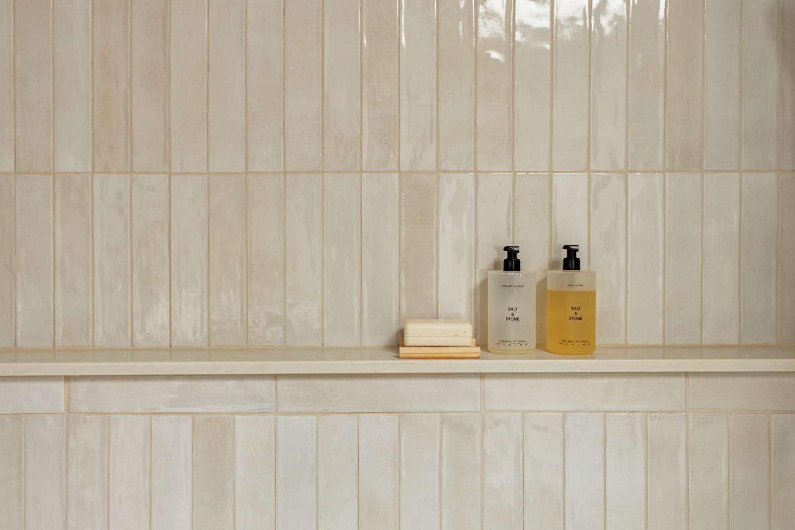 Glazed cream tile wall with a stone ledge running through it, holding two pump bottles and a bar of soap on a wooden dish
