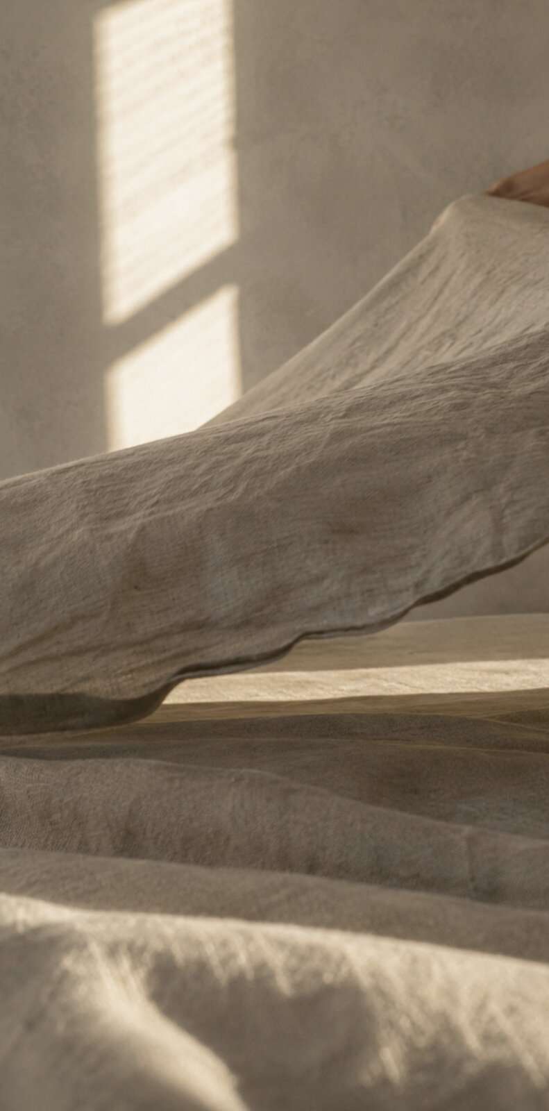 Artist impression — rumpled undyed linen catching low light, with a window's shadow falling across the plaster wall behind