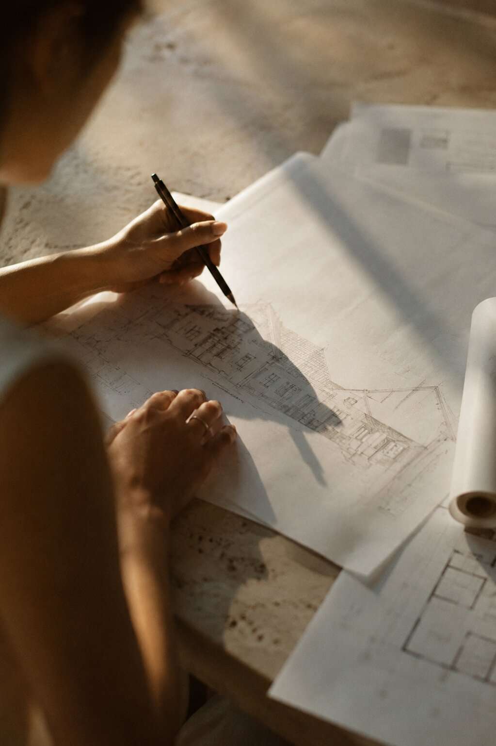 Seen over the shoulder: a hand drawing an elevation in pen on trace paper, with rolled drawings and a floor plan spread across a travertine table in low sun