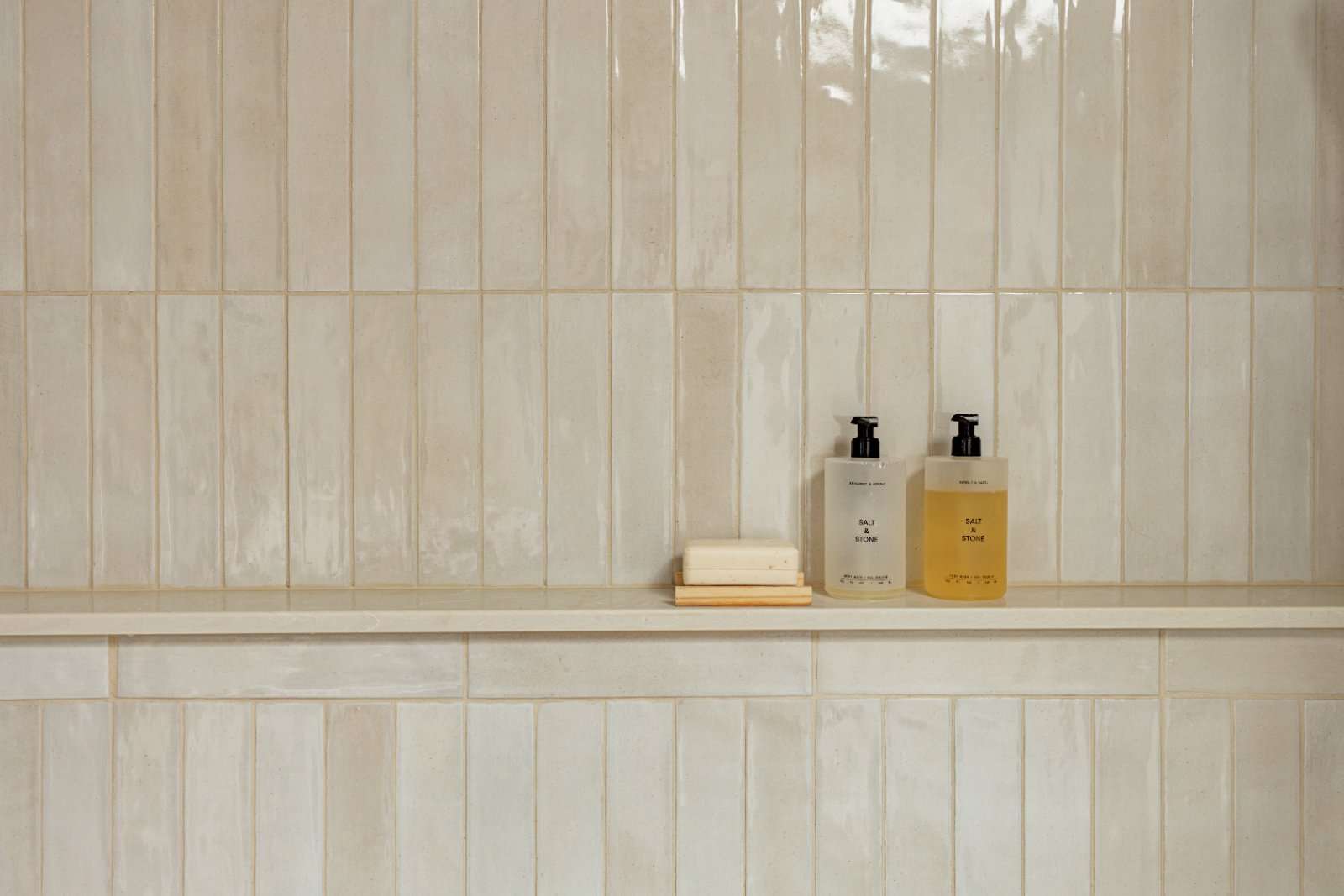 Glazed cream tile wall with a stone ledge running through it, holding two pump bottles and a bar of soap on a wooden dish
